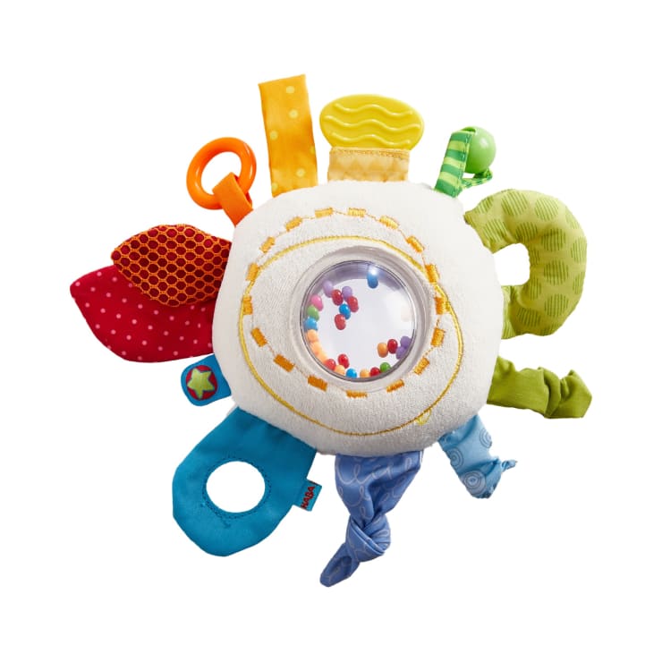Ein rundes, mehrfarbiges Plüsch-Babyspielzeug mit verschiedenen strukturierten Stofflaschen, einem durchsichtigen Kunststofffenster, das kleine farbige Perlen enthält, und mehreren angebrachten Schlaufen und Anhängern, dargestellt auf weißem Hintergrund.