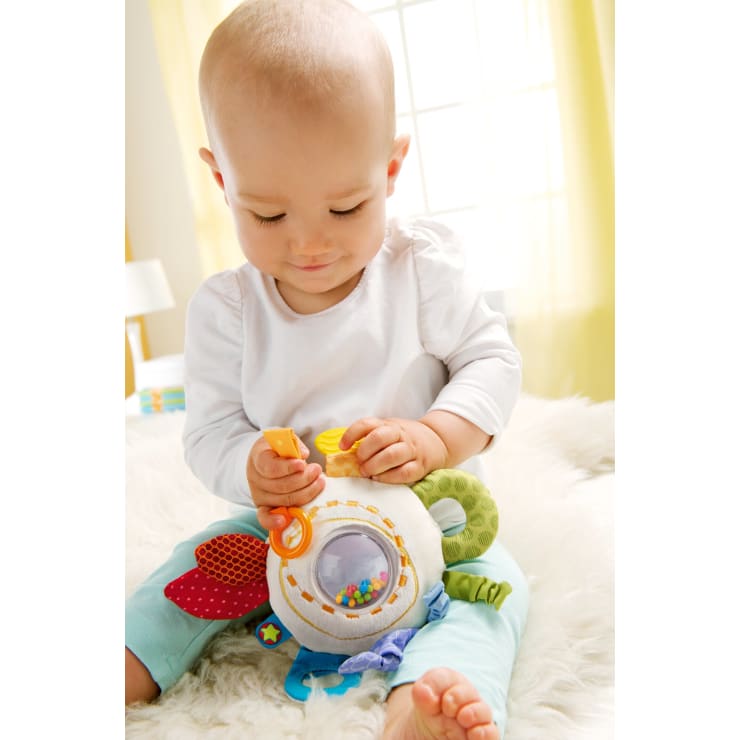 Ein Baby sitzt auf einer weichen, weißen Oberfläche und spielt mit einem bunten, runden Plüschtier, das verschiedene Strukturen, Bänder und einen durchsichtigen Bereich mit kleinen Perlen im Inneren aufweist. Der Hintergrund ist sanft mit natürlichem Licht aus einem Fenster beleuchtet.