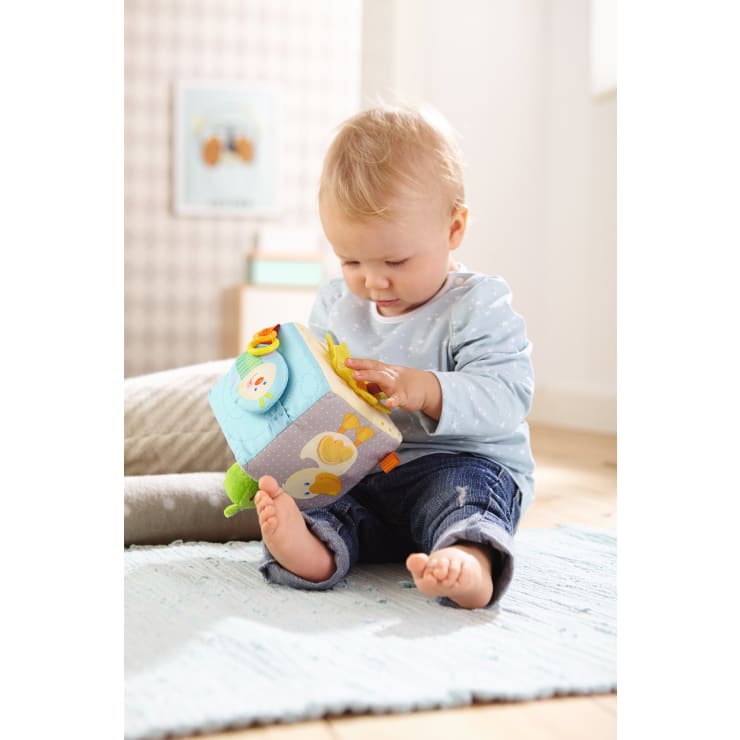 Ein Baby sitzt auf einem hellblauen Teppich und spielt mit einem weichen, bunten Activity Cube. Das Baby trägt ein hellblaues Hemd und Jeans, hat nackte Füße und scheint sich auf das Spielzeug zu konzentrieren. Die Umgebung ist ein helles, gemütliches Zimmer.