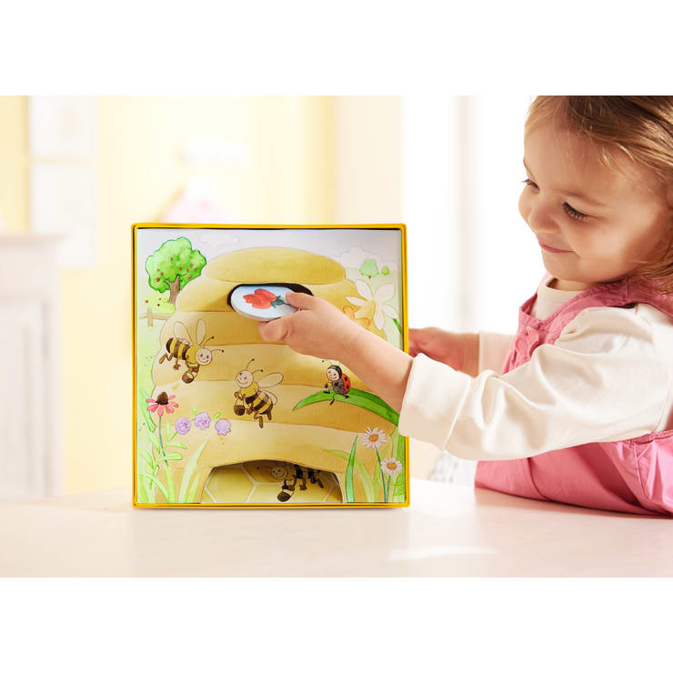 Ein kleines Kind legt eine runde Spielmarke mit einem roten Herz auf eine bunt illustrierte Tafel mit Bienen, Blumen und einem Bienenstock. Das Kind lächelt und trägt ein hellrosa langärmeliges Hemd.
