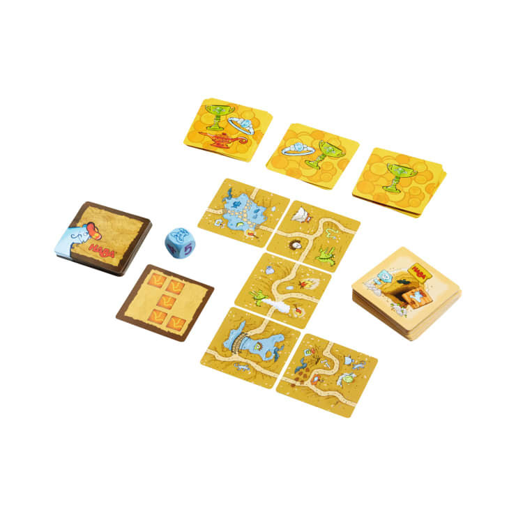 Ein Brettspielset mit einem blauen Würfel, quadratischen, illustrierten Spielsteinen, die Wege und Wasser darstellen, einem Stapel gelber Karten, einer braunen Anweisungskarte und einem hölzernen Aufbewahrungsbehälter, alles auf weißem Hintergrund.
