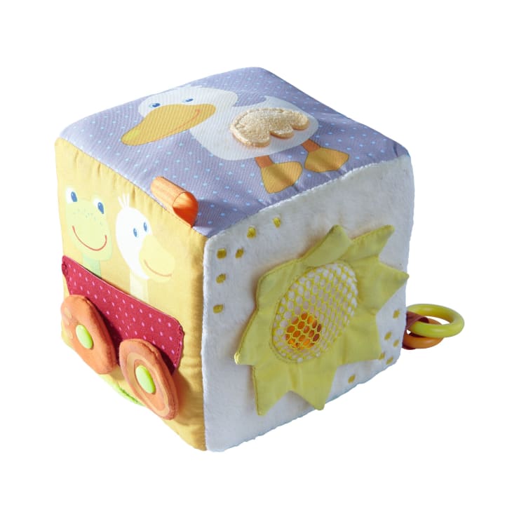 Ein weicher, farbenfroher Stoffspielzeugblock mit Tierillustrationen, Stofftexturen, einer gelben Blume und zwei rosa Rädern, mit Plastikringen an einer Seite, auf weißem Hintergrund.
