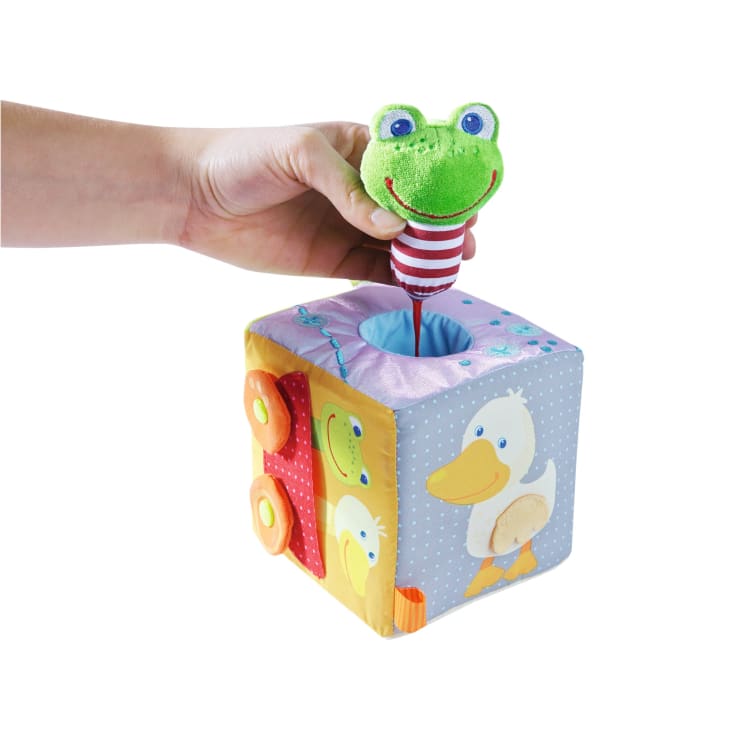 Eine Hand zieht eine grüne Froschpuppe mit einem rot-weiß gestreiften Körper aus einem weichen, bunten Würfelspielzeug, das mit Enten- und Autoillustrationen verziert ist. Der Hintergrund ist einfarbig weiß.