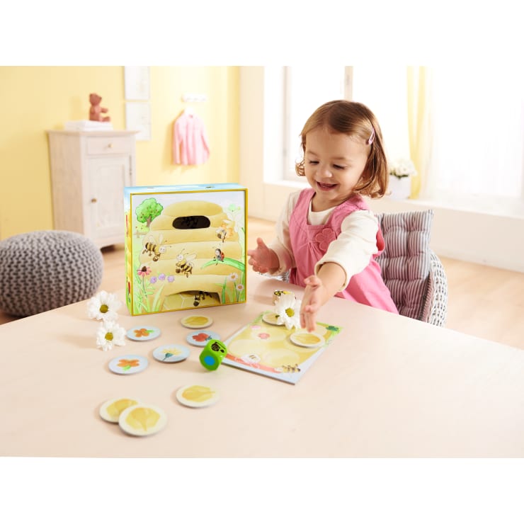Ein kleines Kind in einem rosafarbenen Kleid sitzt an einem Tisch und spielt ein Brettspiel mit runden Spielsteinen und einer Schachtel mit Bienen und einem Bienenstock. Weiße Gänseblümchen stehen auf dem Tisch in einem hellen, luftigen Raum.