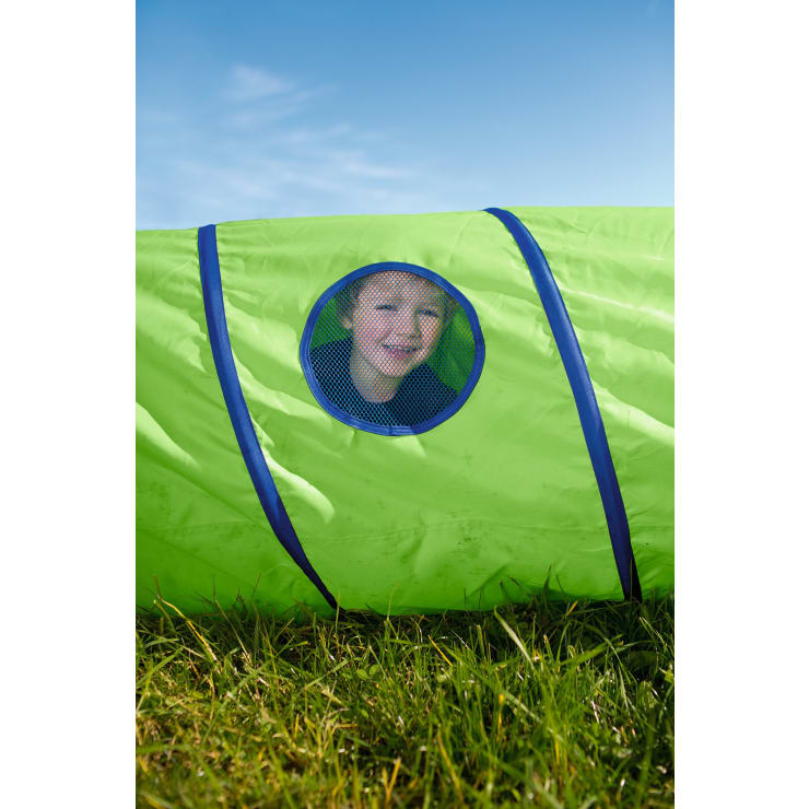 Ein Kind blickt durch ein rundes Netzfenster aus einem hellgrünen Spieltunnel, der auf einer Wiese unter einem klaren blauen Himmel steht.