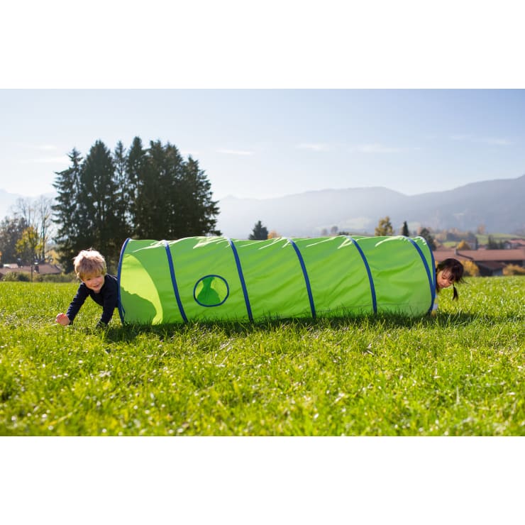 Zwei Kinder krabbeln draußen auf dem Gras und kommen an beiden Enden eines hellgrünen Spieltunnels heraus. Im Hintergrund sind Bäume, Häuser und Berge unter einem klaren blauen Himmel zu sehen.