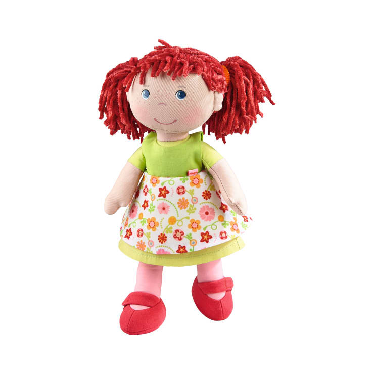 Eine weiche Stoffpuppe mit rotem Garnhaar in Zöpfen, blauen Augen und einem aufgestickten Lächeln, die ein grünes Oberteil, ein weißes Kleid mit Blumenmuster und rote Schuhe trägt und vor einem weißen Hintergrund steht.