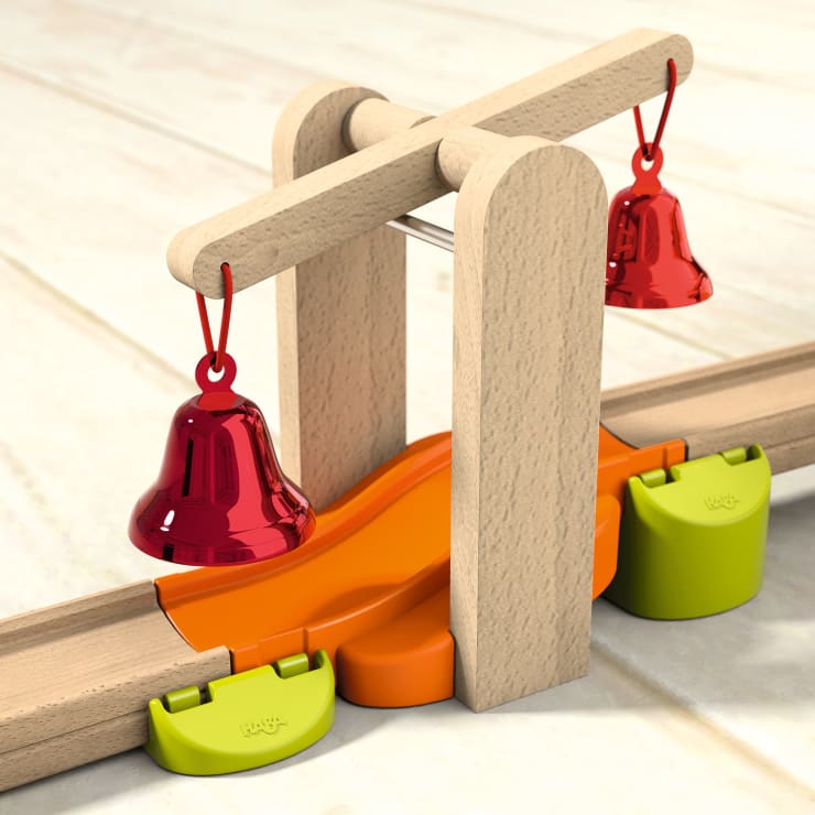 Eine hölzerne Spielzeugbrücke mit zwei roten Glocken, die von beiden Seiten herabhängen, einer orangefarbenen Rampe darunter und grünen Verbindungsstücken, die sie mit hölzernen Eisenbahnschienen auf einer hellen Oberfläche verbinden.