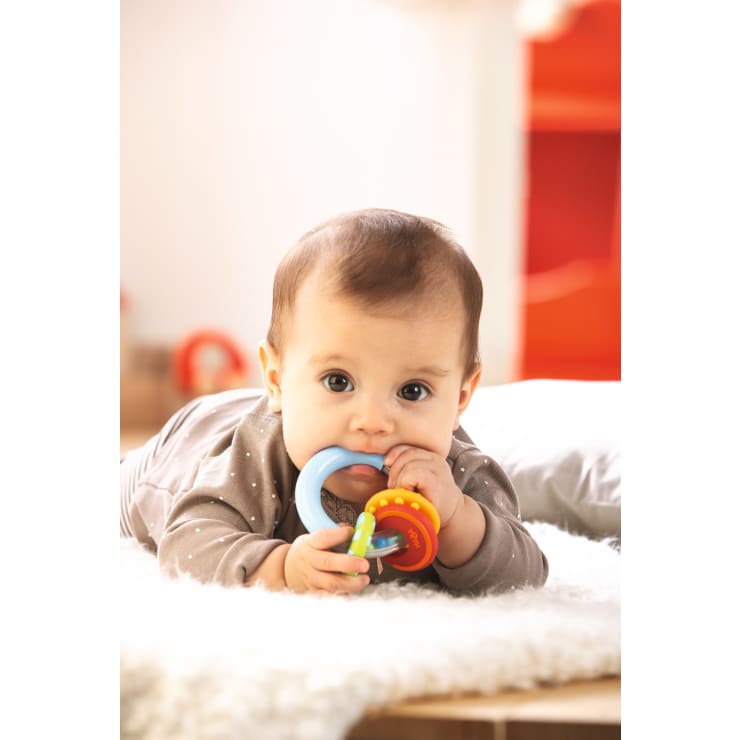 Ein Baby in einem grauen gepunkteten Outfit liegt auf dem Bauch auf einer weichen, weißen Unterlage und kaut auf einem bunten Beißspielzeug aus Plastik mit blauen und roten Ringen.