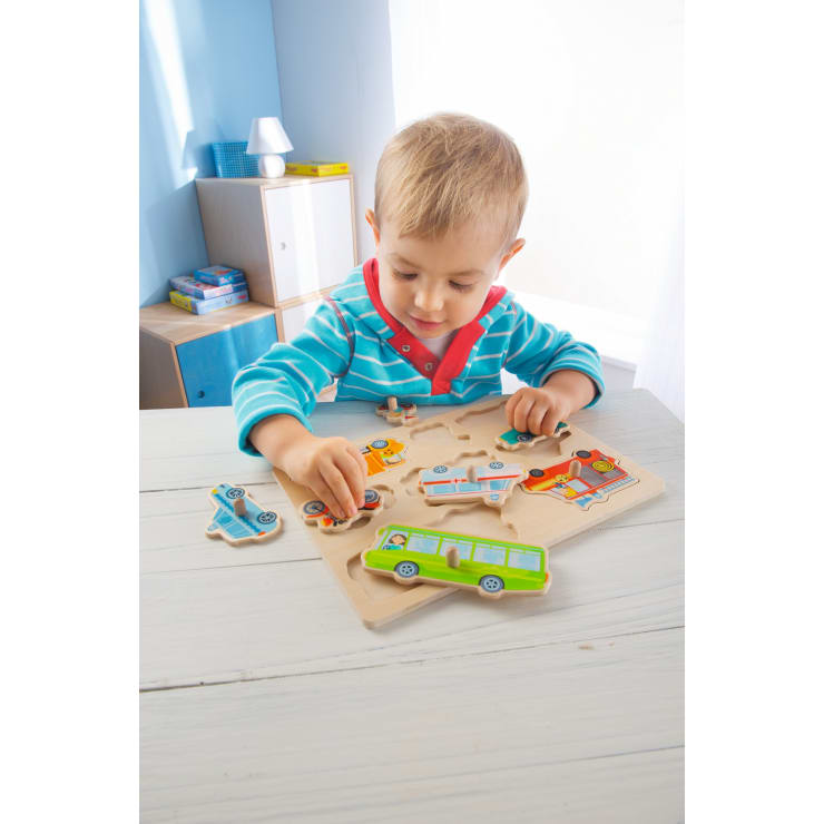 Ein kleines Kind mit blondem Haar und einem blau gestreiften Hemd sitzt an einem weißen Tisch und setzt in einem hellen Raum mit Tageslicht ein Holzpuzzle mit Teilen in Form von Fahrzeugen zusammen.
