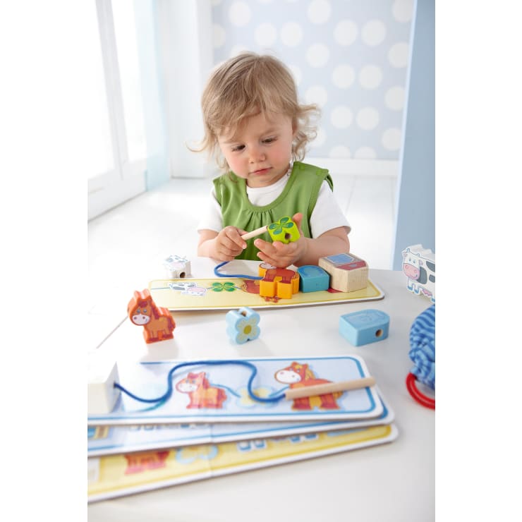 Ein kleines Kind mit blondem Haar sitzt an einem weißen Tisch und spielt mit buntem Holzspielzeug und Puzzleteilen. Das Kind ist konzentriert und hält ein grünes Teil in der Hand, während andere Formen und Tierfiguren auf dem Tisch verstreut sind.