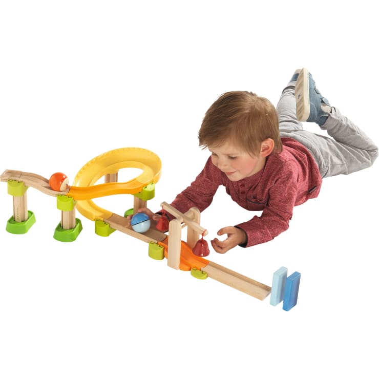 Ein kleines Kind in einem roten Hemd und einer blauen Hose liegt auf dem Boden und spielt mit einer hölzernen Murmelbahn mit bunten Rampen, Blöcken und Murmeln.