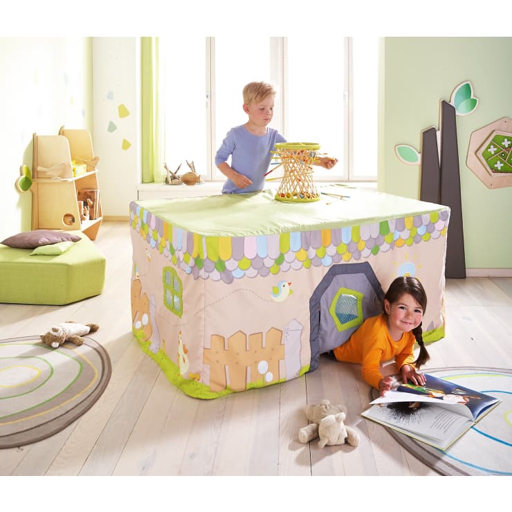 Ein kleiner Junge und ein Mädchen spielen in einem hell erleuchteten Kinderzimmer. Das Mädchen krabbelt aus einem Spielhaus, das aus einer verzierten Tischdecke besteht, während der Junge dahinter steht. Kuscheltiere und Bücher liegen auf dem hellen Holzboden.