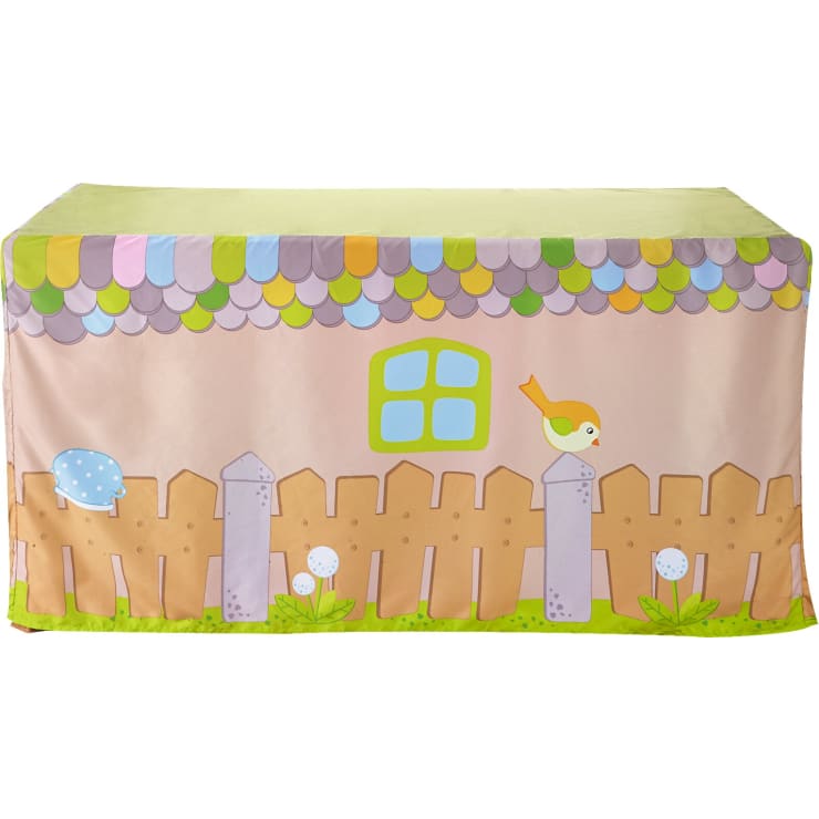Ein rechteckiger Tisch ist mit einem Stoff bedeckt, der wie ein Haus bedruckt ist und einen Lattenzaun, ein Fenster, bunte Dachziegel, einen Vogel auf einem Zaunpfahl, Pilze und Gras zeigt.