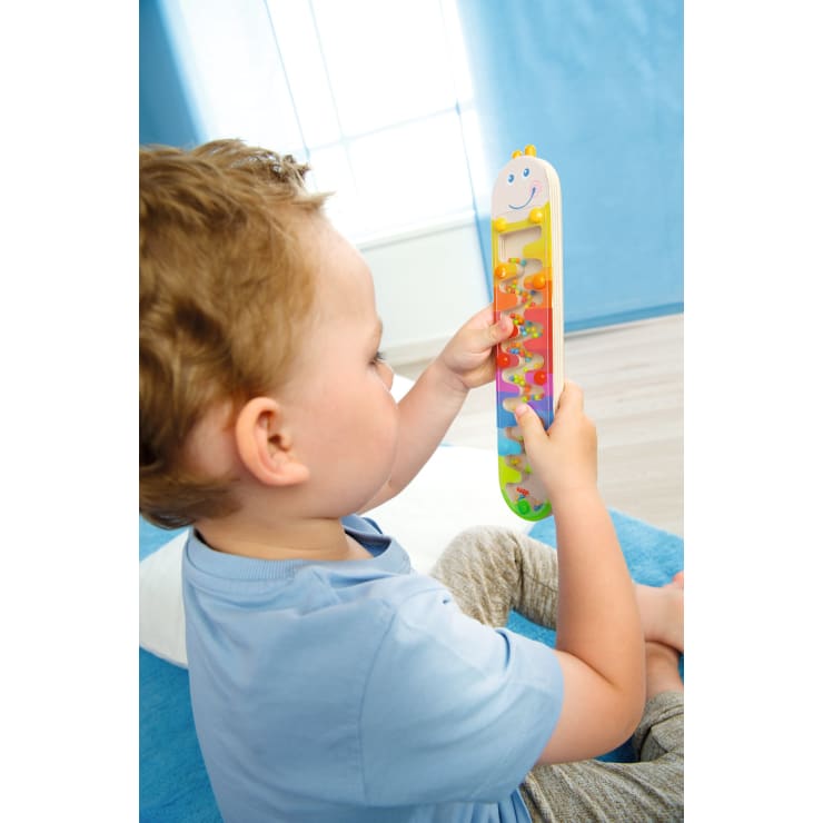 Ein kleines Kind mit hellbraunem Haar und blauem Hemd sitzt auf einem Bett und untersucht in einem hellen Zimmer ein buntes vertikales Spielzeug mit Schiebeperlen.