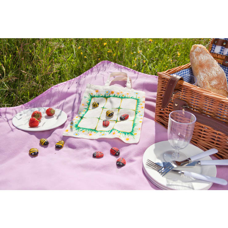 Ein Picknick auf einer Lavendeldecke mit einem Weidenkorb, Brot, Tellern, Besteck, Gläsern, Erdbeeren auf einem Teller und einer Serviette, die als Tic-Tac-Toe-Brett mit aufgemalten Steinbienen und Marienkäfern dient.