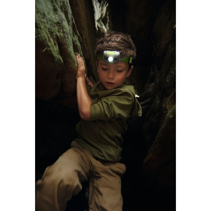 Ein kleiner Junge mit Stirnlampe und Outdoor-Kleidung klettert zwischen großen Felsen in einem engen, schwach beleuchteten Raum. Er stützt sich mit der Hand auf dem Felsen ab und scheint sich darauf zu konzentrieren, das Terrain zu durchqueren.