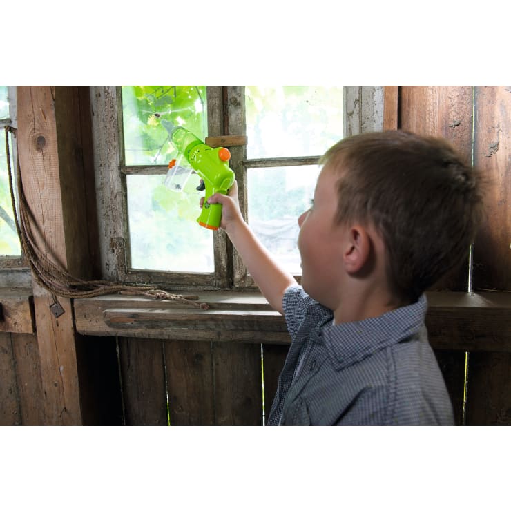 Ein Junge in einem kurzärmeligen, karierten Hemd richtet eine grüne Plastikwasserpistole auf ein altes Holzfenster in einem rustikalen Raum mit Holzwänden.