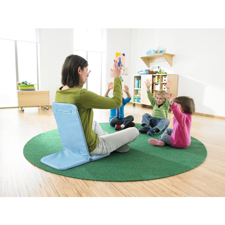 Ein Erwachsener sitzt auf einer grünen Matte vor vier Kindern, die ebenfalls mit erhobenen Armen auf der Matte sitzen; die Umgebung scheint ein helles, organisiertes Klassenzimmer mit Regalen und Unterrichtsmaterialien zu sein.