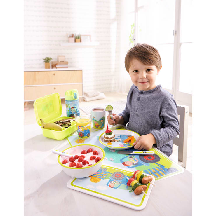 Ein Junge sitzt an einem Tisch mit einem bunten Tischset und isst eine Mahlzeit, die eine Obstschale, ein Sandwich, geschnittenes Gemüse, einen Spieß und ein Getränk umfasst. Auf dem Tisch stehen eine Brotdose und eine Wasserflasche.