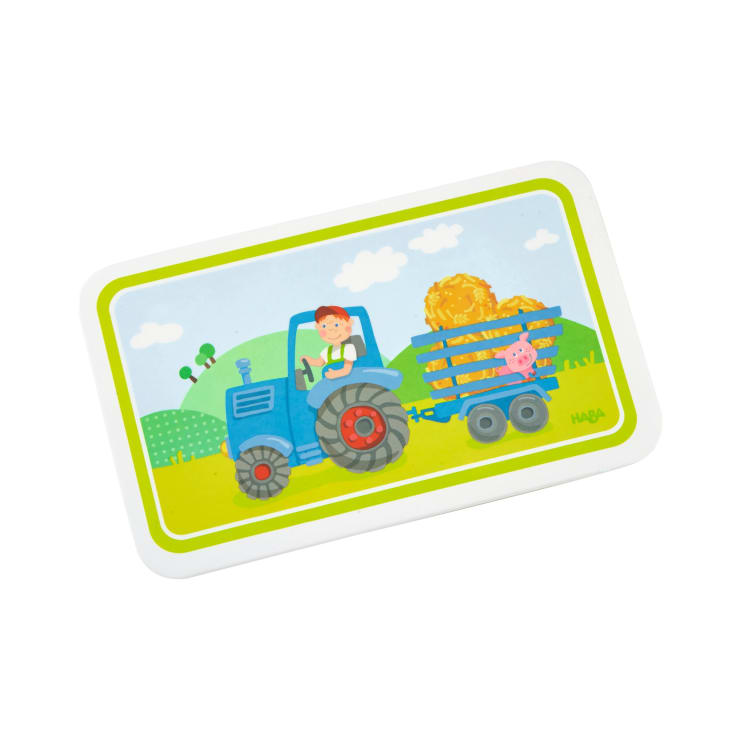 Eine illustrierte Karte zeigt ein Kind, das einen blauen Traktor fährt, der einen Anhänger mit Heuballen und einem rosa Schwein zieht, inmitten einer grünen Landschaft mit Hügeln, Wolken und einem gelben Rand.