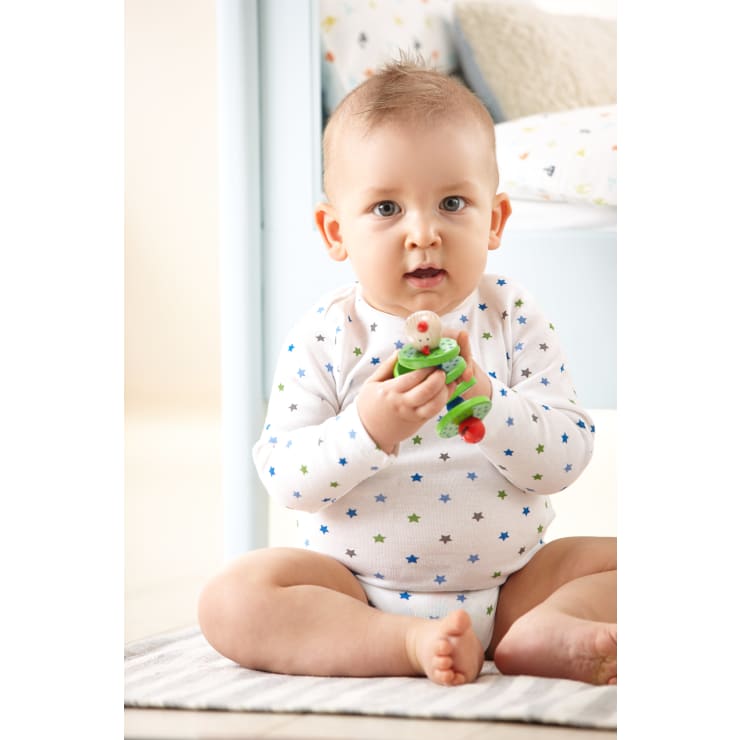 Ein Baby, das einen weißen Strampler mit bunten Sternen trägt, sitzt auf einer weichen Matte und hält ein grünes und rotes Spielzeug. Das Baby hat helle Haut und kurze Haare. Im Hintergrund ist ein Kinderbett mit gemustertem Bettzeug zu sehen.