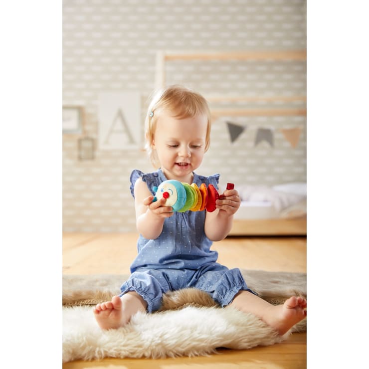 Ein Kleinkind mit blondem Haar sitzt auf einem weichen Teppich in einem Kinderzimmer, trägt ein blaues Outfit und hält ein buntes Holzspielzeug, das es interessiert betrachtet. Das Zimmer hat ein Kinderbett und Wanddekorationen im Hintergrund.