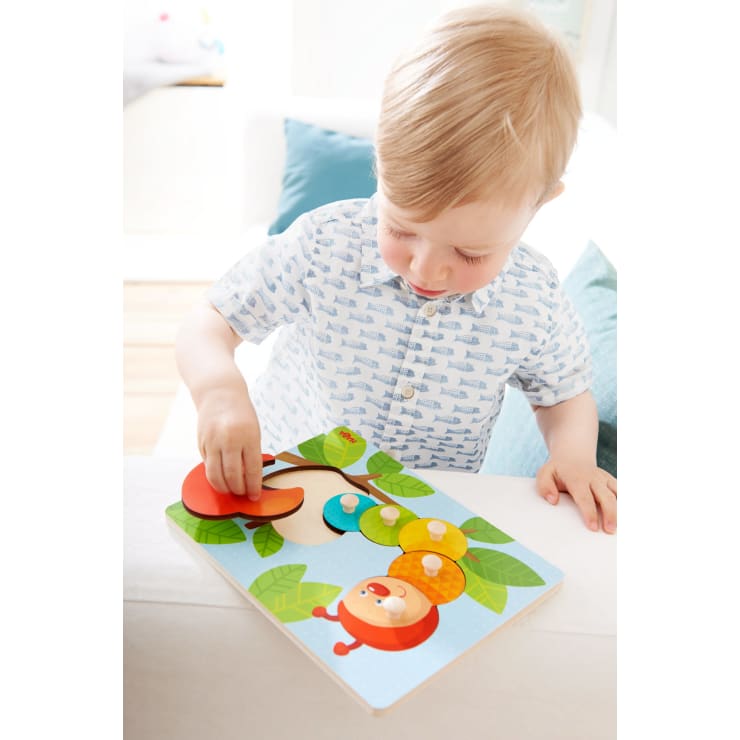 Ein kleines Kind mit blondem Haar und einem gemusterten Hemd legt ein rundes Teil in ein buntes Holzpuzzle mit einem Marienkäfer und Blättern auf einem Tisch im Haus.