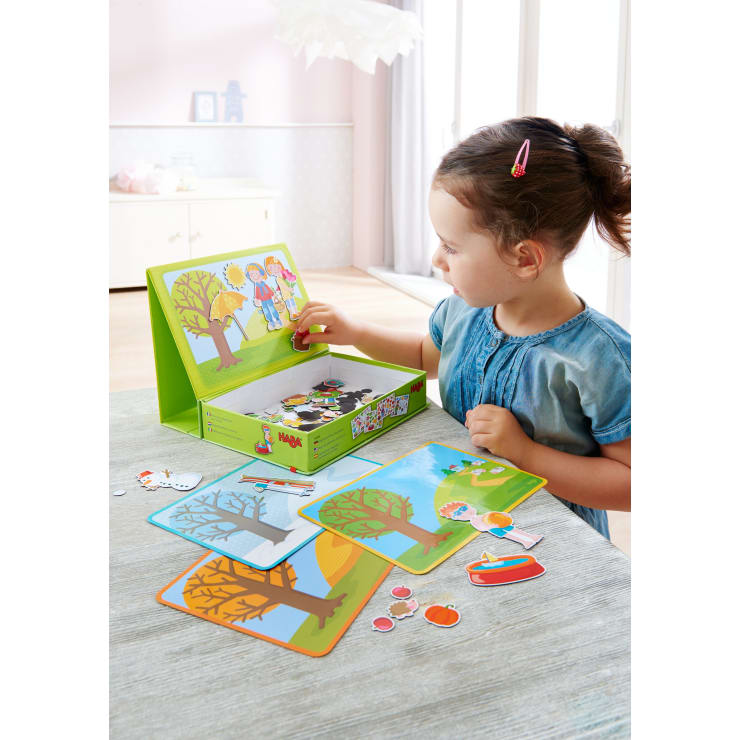 Ein junges Mädchen sitzt an einem Tisch und spielt mit einem magnetischen Brettspiel, das illustrierte Karten, Magnete von Menschen, Bäumen und Gegenständen enthält. Der Raum ist hell mit natürlichem Licht, das durch große Fenster fällt.