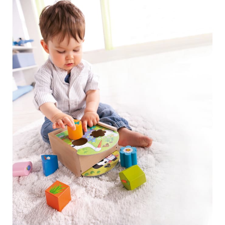 Ein kleines Kind sitzt auf einem weichen, weißen Teppich und spielt mit einem hölzernen Sortierspielzeug und steckt einen farbigen Block in eines der Löcher. Andere bunte Blöcke liegen in der Nähe verstreut.