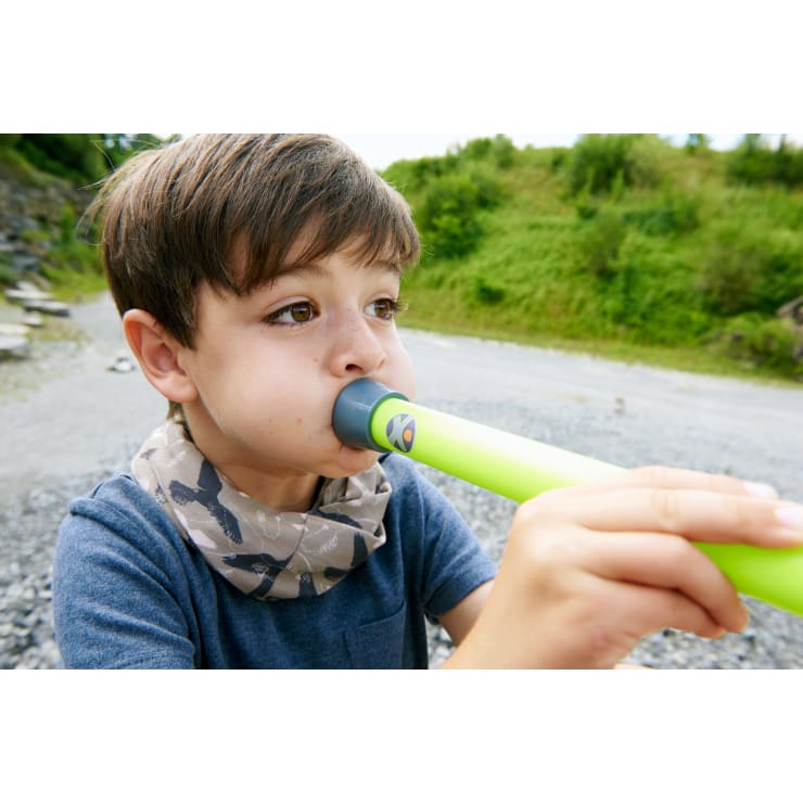 Ein kleiner Junge benutzt im Freien einen leuchtend grünen Atemschlauch. Er trägt ein blaues Hemd und einen grauen Schal mit dunklen Mustern. Im Hintergrund sind Grünflächen, Felsen und ein Kiesweg zu sehen.
