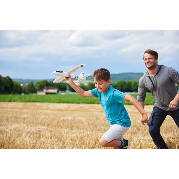 Ein Junge in einem blauen Hemd rennt mit einem Spielzeugflugzeug in der Hand durch ein Strohfeld, während ein erwachsener Mann hinter ihm steht. Der Himmel ist teilweise bewölkt und im Hintergrund sind Bäume und Gebäude zu sehen.
