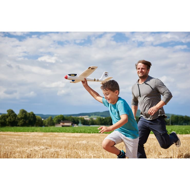 Ein Junge läuft mit einem Modellflugzeug in der Hand durch ein Feld, während ein erwachsener Mann hinter ihm herläuft. Der Himmel ist teilweise bewölkt, und im Hintergrund sind Bäume und ein Haus zu sehen. Beide scheinen sich an der Aktivität im Freien zu erfreuen.