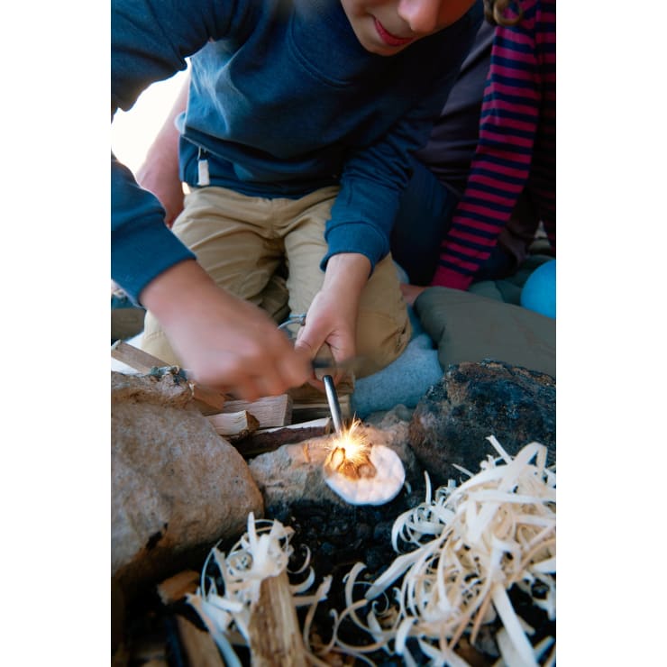 Eine Person versucht, mit einem Feueranzünder Funken über Anzündholz und kleine Holzspäne zu schlagen, um ein Feuer im Freien zu entfachen. In der Nähe sind große Felsen und eine weitere Person in gestreifter Kleidung zu sehen.
