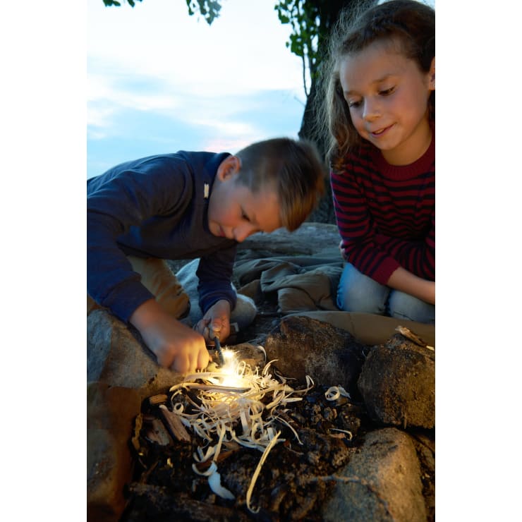 Ein Junge zündet mit einem Feueranzünder Holzspäne in einem Lagerfeuerring an, während ein Mädchen in der Nähe sitzt und zuschaut. Beide Kinder sind im Freien, und es scheint Abend zu sein.