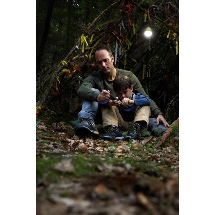 Ein Erwachsener und ein Kind sitzen auf dem Waldboden und schnitzen gemeinsam mit einem Messer einen Stock. Hinter ihnen steht ein behelfsmäßiger Unterstand, der mit gelben, weißen und roten Bändern geschmückt ist. Abgefallene Blätter bedecken den Boden.