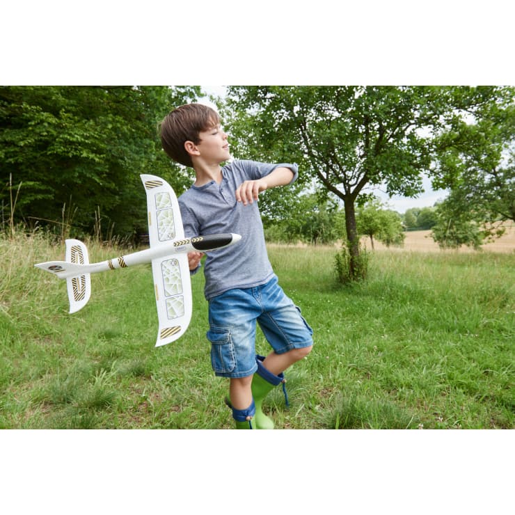 Ein Junge in einem grauen Hemd, Jeans-Shorts und grünen Stiefeln steht im Gras und hält ein großes weißes Spielzeugflugzeug in der Hand, mit Bäumen und einem Feld im Hintergrund.