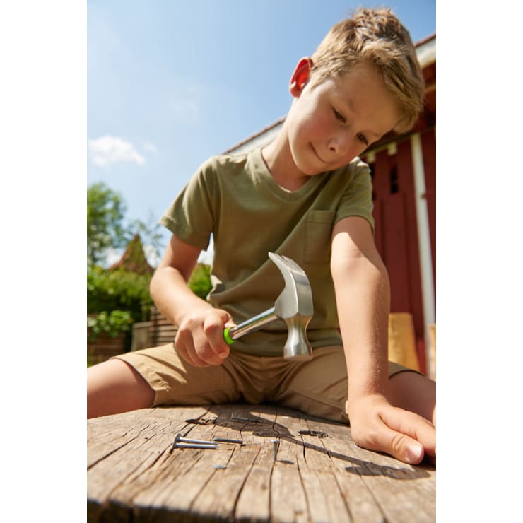 Ein Junge in grünem T-Shirt und Shorts sitzt an einem sonnigen Tag draußen auf einer Holzfläche, hält einen Hammer in der Hand und bereitet sich darauf vor, einen Nagel in das Holz zu schlagen.