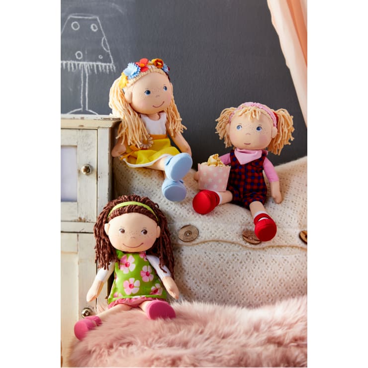 Drei Stoffpuppen mit Garnhaaren sitzen auf einer cremefarbenen Strickcouch; eine trägt ein gelbes Kleid mit einem Blumenstirnband, eine hat rote Schuhe mit Latzhose und eine Geschenktüte, und eine hat ein grünes Blumenkleid und ein Stirnband.