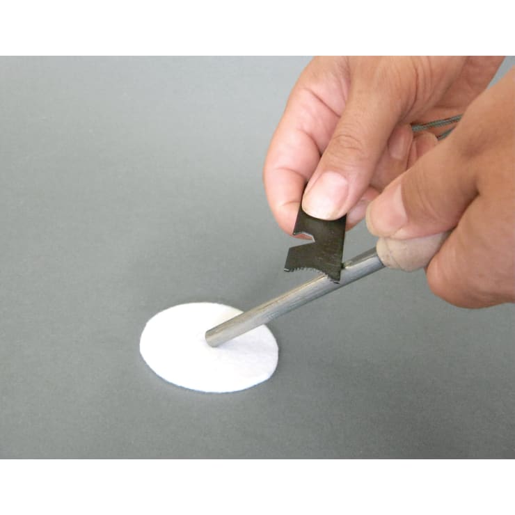 Eine Person hält einen zylindrischen Metallstab über ein weißes, rundes Objekt auf einer grauen Fläche und schabt es mit einem kleinen Metallwerkzeug.