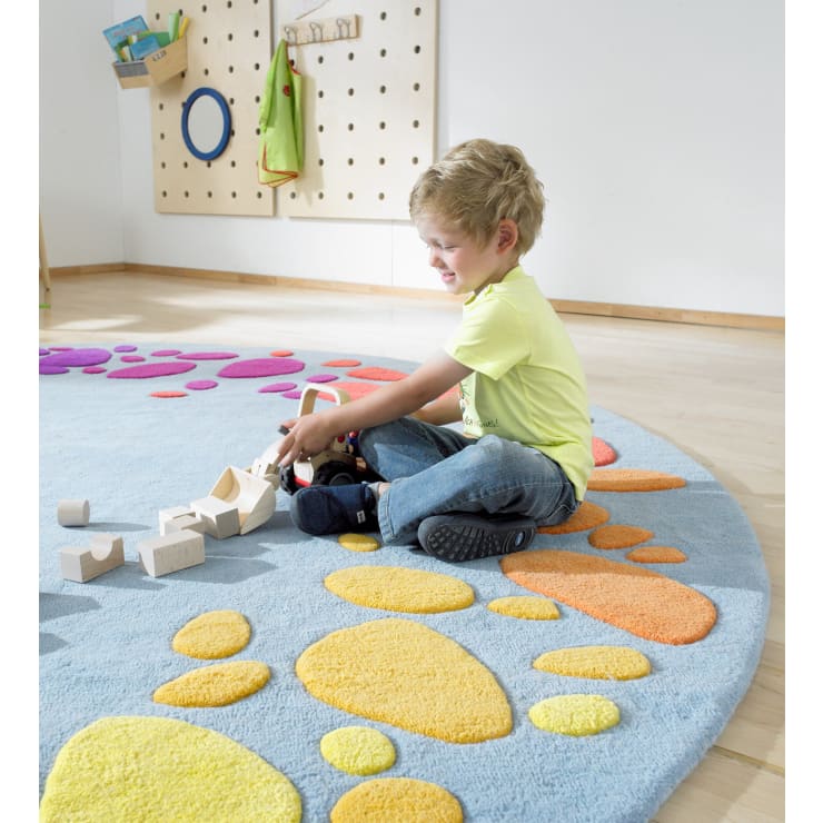 Ein kleines Kind sitzt auf einem blauen Teppich mit orangefarbenen und gelben Formen und spielt mit Holzklötzen. Der Raum hat einen hellen Holzboden und eine Wand mit Haken, an denen kleine Gegenstände hängen.