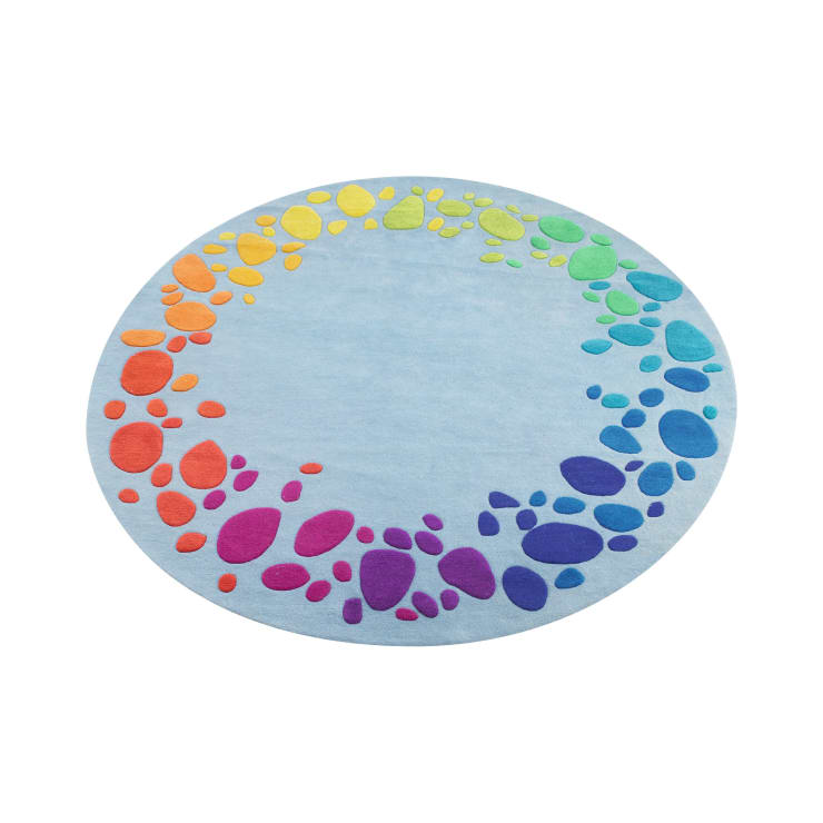 Ein runder, hellblauer Teppich mit mehrfarbigen, kieselsteinähnlichen Formen, die in einem kreisförmigen Muster entlang der Kante angeordnet sind und durch die Farben des Regenbogens verlaufen.