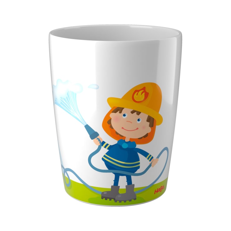 Eine weiße Tasse mit der Illustration eines lächelnden Kindes, das eine Feuerwehruniform und einen Helm trägt, Wasser aus einem Schlauch spritzt und auf grünem Gras steht.