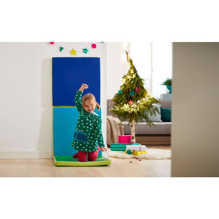Ein kleines Kind in einem grün gepunkteten Kleid kniet auf einer Matte und hebt einen Arm in die Luft. Dahinter steht ein kleiner geschmückter Weihnachtsbaum mit Lichtern, Geschenken und einem Sofa in einem hell erleuchteten Raum.