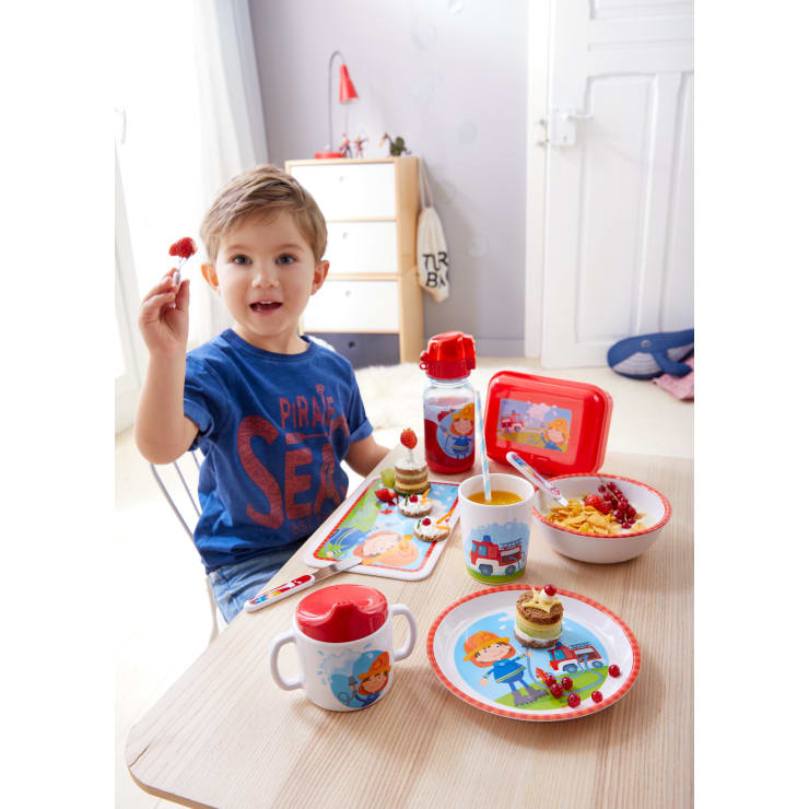 Ein kleiner Junge sitzt an einem kleinen Tisch mit einem thematischen Frühstücksset und hält eine Erdbeere in der Hand. Auf dem Tisch stehen Teller, Tassen, eine Schüssel mit Müsli, ein Getränk, etwas Obst und eine Wasserflasche, alles mit bunten Cartoon-Motiven.