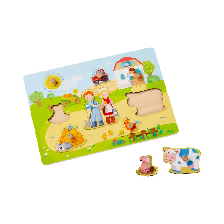 Ein Holzpuzzle zum Thema Bauernhof mit drei herausnehmbaren Teilen - einer Kuh, einem Schwein auf einem Heuhaufen und einem Traktor. Das Puzzlebrett zeigt eine Bauernhoflandschaft mit einer Scheune, zwei Menschen, einem Huhn und einer Katze.