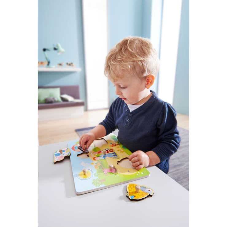 Ein kleines Kind mit blonden Haaren und einem marineblauen Hemd legt ein Teil in ein buntes Holzpuzzle mit Tiermotiven auf einem weißen Tisch in einem hellblauen Zimmer.