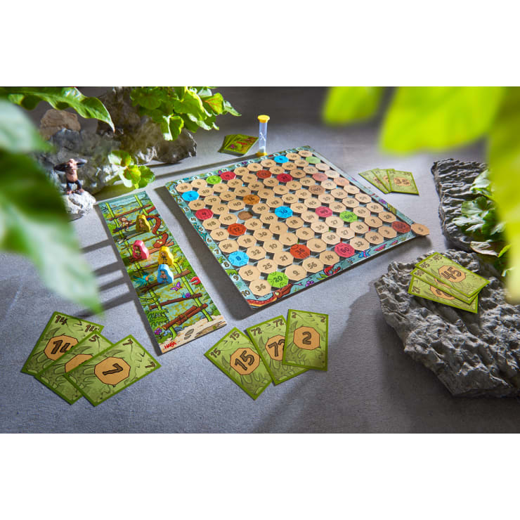 Ein Brettspiel mit einem nummerierten Raster, Holzscheiben, einer Sanduhr, einem farbig illustrierten Seitenbrett und grünen nummerierten Karten wird auf einer Betonfläche mit dekorativen Steinen und Blättern aufgestellt.