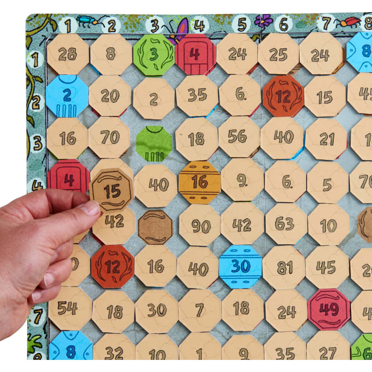 Eine Hand legt ein sechseckiges Plättchen mit der Aufschrift "15" auf ein Spielbrett, das mit nummerierten Plättchen in verschiedenen Farben gefüllt ist, darunter rot, gelb, grün und blau. Das Spielbrett hat nummerierte Felder von 1 bis 81.