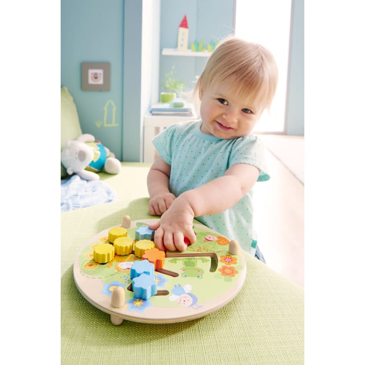 Ein Kleinkind mit hellem Haar und einem hellblauen Polka-Dot-Shirt sitzt an einem Tisch und spielt mit einem bunten Holzpuzzle mit Zahnrädern und Naturillustrationen in einem hell erleuchteten Raum.